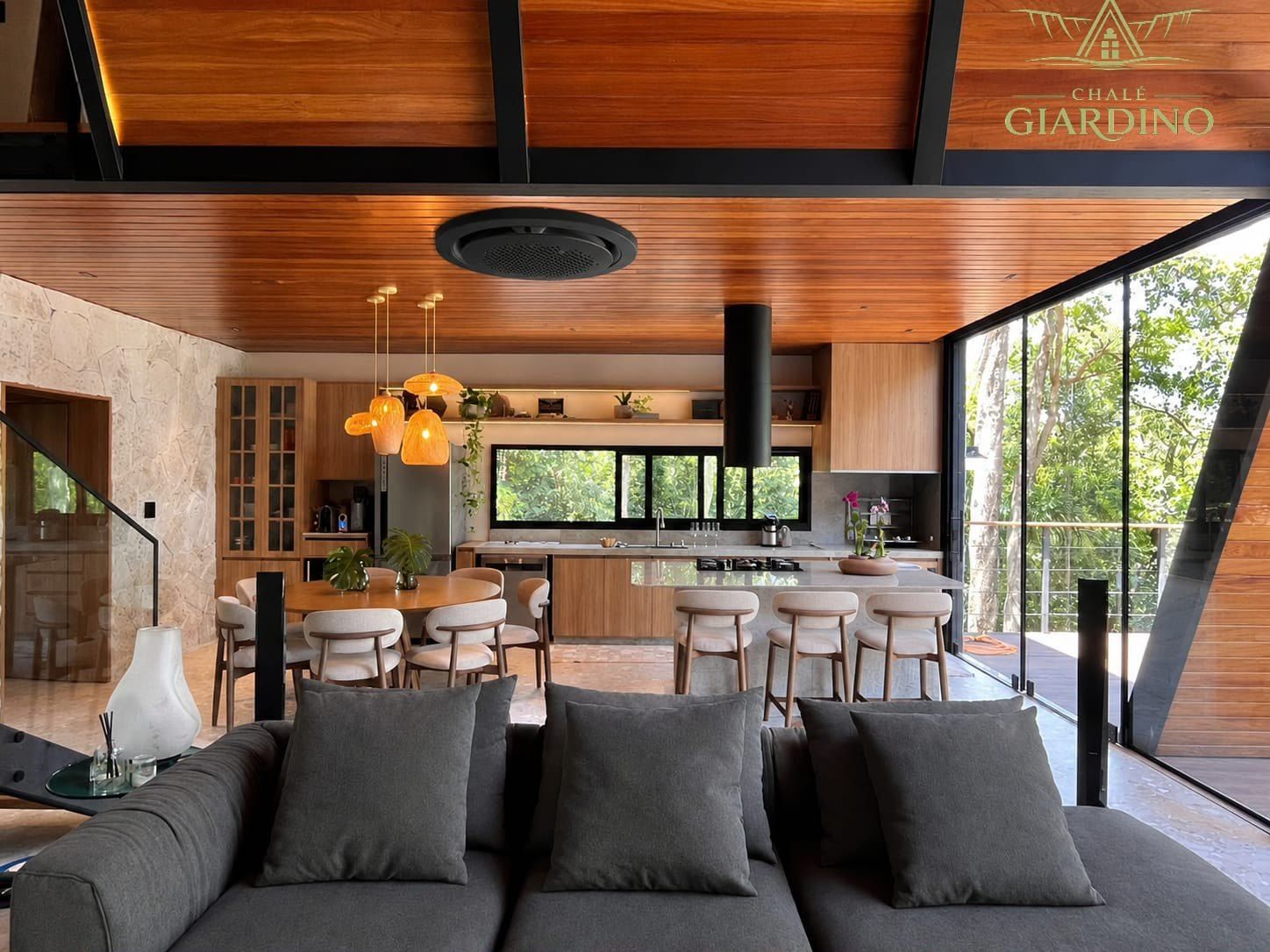 Sala com sofá modular cinza integrada à cozinha e ao jantar, com fachada de vidro para a mata